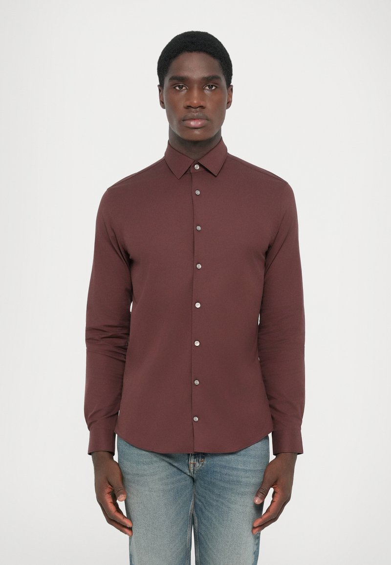 Camisa de manga larga burdeos con cuello abotonado, botones plateados y un corte entallado, combinada con unos vaqueros azul claros. Textura suave.