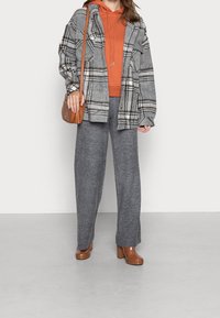 Šedá kostkovaná oversized flanelová bunda, oranžová mikina s kapucí, šedé široké pletené kalhoty, hnědé boty na podpatku a hnědá kabelka přes rameno.