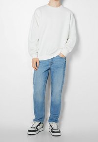 Sudadera blanca de manga larga y cuello redondo, combinada con jeans rectos de mezclilla azul claro y zapatillas negras y blancas.