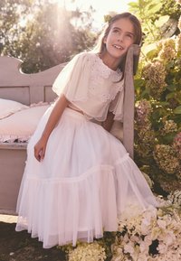 Un conjunto blanco de dos piezas que presenta una blusa de encaje transparente bordada con acentos florales y una falda de tul en capas. Entorno natural con flores.