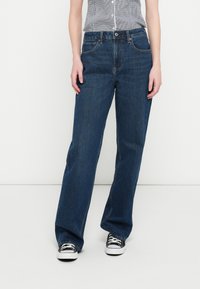 Mørk denim viddeben jeans med h�øj talje, der har et design med fem lommer og diskrete syningsdetaljer. Båret med sorte og hvide sneakers.