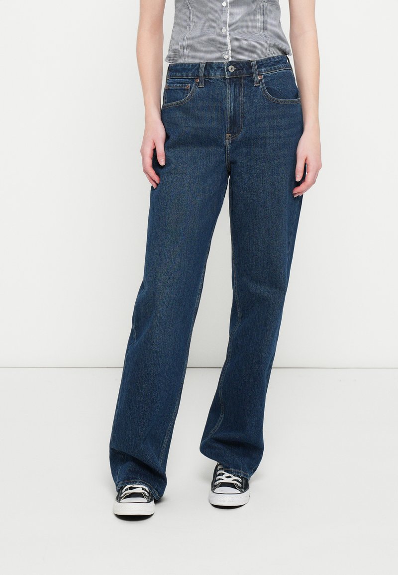 Mørk denim viddeben jeans med høj talje, der har et design med fem lommer og diskrete syningsdetaljer. Båret med sorte og hvide sneakers.