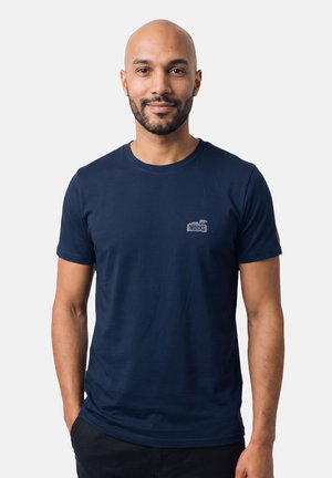 Mann mit Bart und rasiertem Kopf, der ein dunkelblaues T-Shirt mit einem kleinen "WORKS" Logo auf der Brust trägt, vor einem einfarbigen hellen Hintergrund stehend.