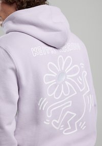 Laventeli huppari, jossa on valkoinen Keith Haringin graafinen tanssiva hahmo ja kukka. Sisältää kiristettävän hupun ja ribbitaskut.