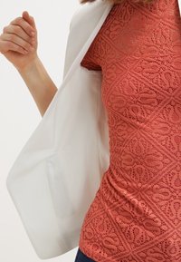 Donna che indossa un top in pizzo corallo a fantasia mentre infila il braccio sinistro nella manica di un blazer bianco.