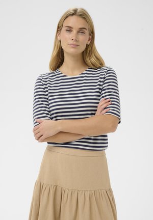 Giovane donna con capelli biondi che indossa una maglietta a righe navy e bianche e una gonna beige, in piedi con le braccia incrociate davanti a uno sfondo neutro.