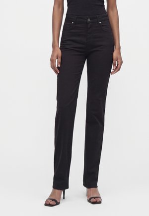 Femme portant un pantalon noir slim taille haute et des sandales noires à talons, debout devant un fond clair uni.