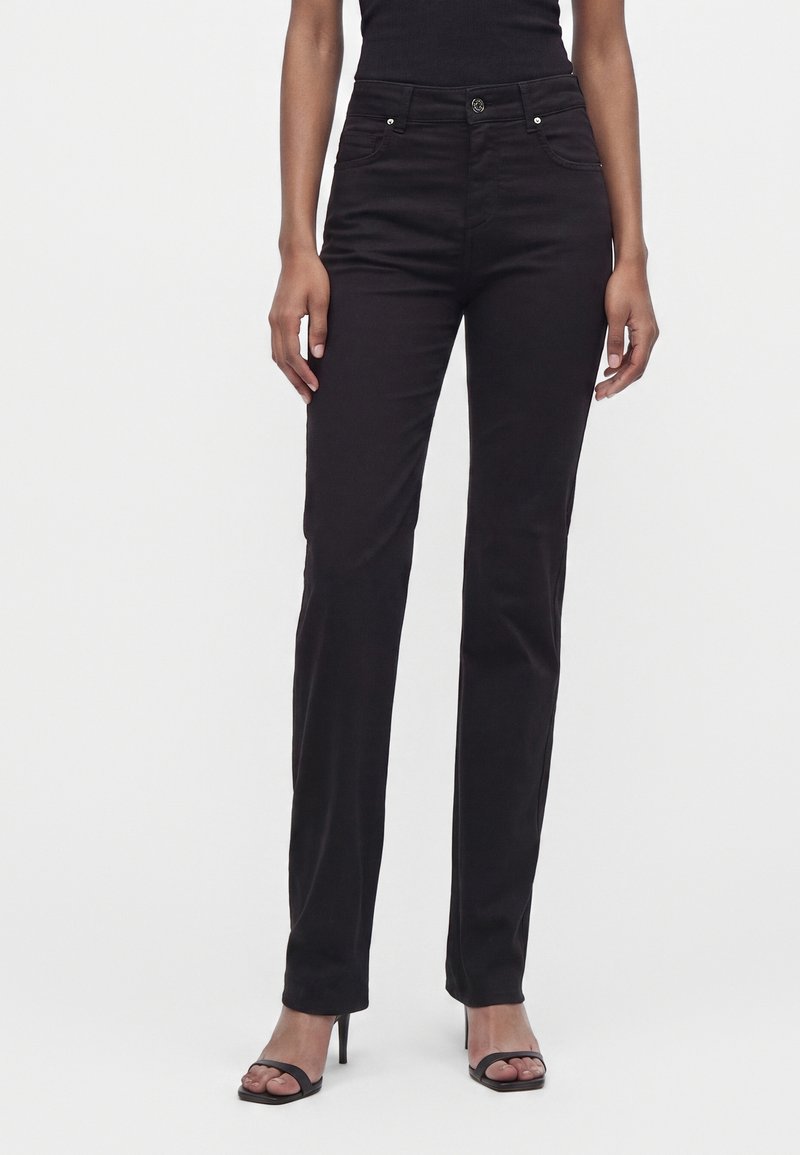 Femme portant un pantalon noir slim taille haute et des sandales noires à talons, debout devant un fond clair uni.