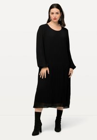Musta pleated-keskitason mekko, jossa on pitkät hihat ja pyöreä kaula-aukko. Yhdistetty mustiin nilkkureihin. Sujuva, virtaava kankaan rakenne.