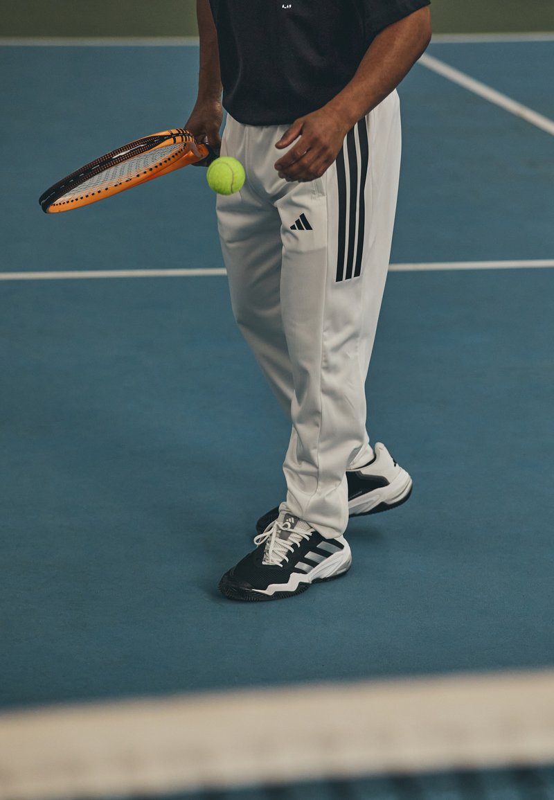 Jugador de tenis sosteniendo una raqueta naranja, vistiendo zapatillas negras con acentos blancos y pantalones blancos con franjas negras a los lados. Pelota de tenis amarilla visible.