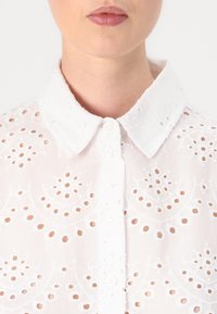 White embroidered blouse featuring a collared neckline, button down front, and intricate cut-out patterns throughout the fabric.