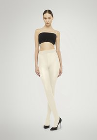 Leggings beige realizzati in un materiale liscio, abbinati a un top bandeau nero e tacchi a spillo neri, su uno sfondo neutro.