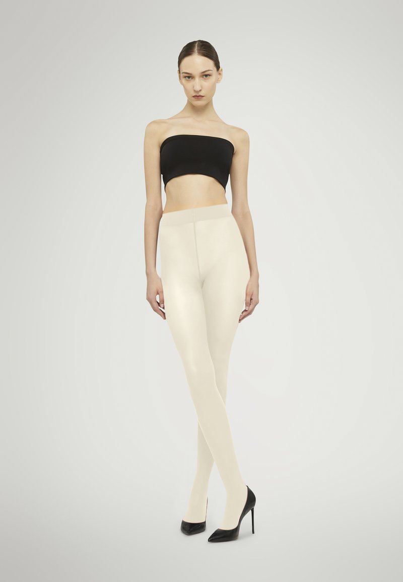 Leggings beige realizzati in un materiale liscio, abbinati a un top bandeau nero e tacchi a spillo neri, su uno sfondo neutro.
