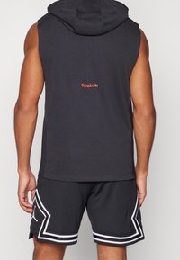 Hombre con una sudadera sin mangas negra de Reebok y pantalones cortos de baloncesto negros con ribetes blancos, visto desde atrás sobre un fondo liso.