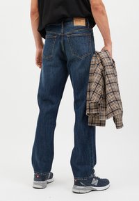 Pantalones vaqueros de mezclilla azul oscuro con corte de pierna recta, bolsillos traseros y una etiqueta marrón en la cinturilla, combinados con una camisa a cuadros colocada sobre una mano.
