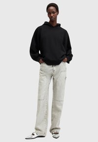 Sudadera negra con capucha y corte holgado, combinada con jeans de patas anchas de color gris claro que tienen bolsillos frontales y detalles desgastados. Zapatos plateados de punta afilada.