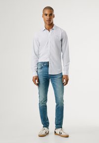 Camisa de botón ajustada de rayas azul claro, con mangas largas. Combinada con jeans azul desgastado y zapatillas blancas con detalles en negro.