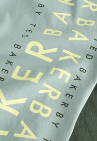 Baumwollstoff in Hellblau mit großen gelben und schwarzen Buchstaben: "TED BAKER" auffällig in wechselnden Ausrichtungen und Stilen präsentiert.