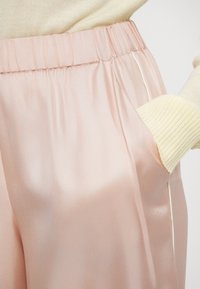 Roosad satiinist püksid, millel on elastne vöökoht, küljetaskud ja sile, läikiv tekstuur. Komplekt on kombineeritud hele kollase soonikkoega kampsuniga.