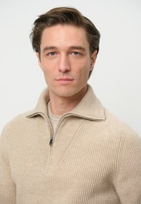 Suéter de punto beige con cuello con cremallera y textura de canalé. Presenta un diseño en forma de V en la parte delantera. Llevado sobre una camisa de color claro.