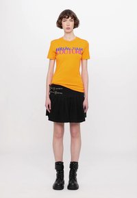 Oranžové tričko s textem "VERSACE JEANS COUTURE", v kombinaci s černou skládací sukní se sponovými akcenty a černými kotníkovými botami.