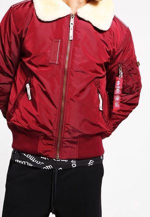 Veste bomber rouge avec col polaire blanc, fermeture éclair devant, poches latérales et une poche zippée sur la manche avec une étiquette rouge "Remove Before Flight".