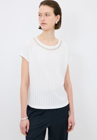 Top blanco de manga corta con un patrón texturizado, cuello redondo decorado con cuentas similares a perlas, acompañado de pantalones oscuros.
