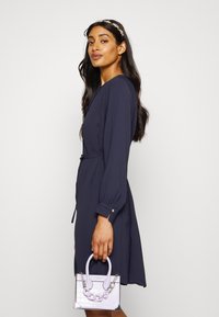 Vestido azul marinho com mangas longas, cintura ajustada e textura suave. Segurando uma mala lilás com acabamento texturizado e detalhes em corrente.