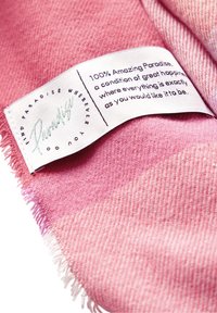 Rosa gewebter Stoff mit einem ausgefransten Rand, versehen mit einem weißen Stoffetikett, auf dem schwarzer Text und ein stilisiertes grünes Logo mit der Aufschrift "Paradise" zu sehen sind.