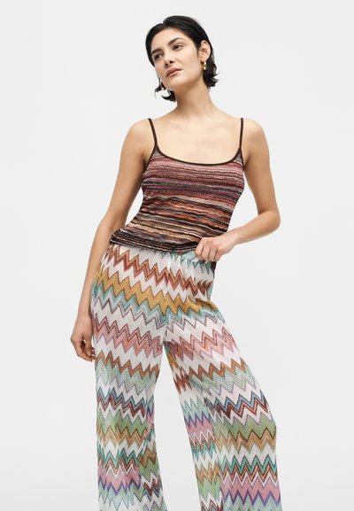 Νεαρή γυναίκα ποζάρει με πολύχρωμο παντελόνι φαρδιάς γραμμής με ζιγκ ζαγκ μοτίβο και αμάνικο ριγέ τοπ σε απλό λευκό φόντο.