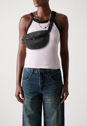 Žena v bílém tílku, modrých džínách, s černou kabelkou přes rameno a stříbrným řetízkovým náhrdelníkem na jednobarevném pozadí.