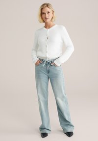 Weißer gestrickter Cardigan mit Rundhalsausschnitt und Knopfverschluss, kombiniert mit hellblauen weiten Jeans und schwarzen spitzen Schuhen.