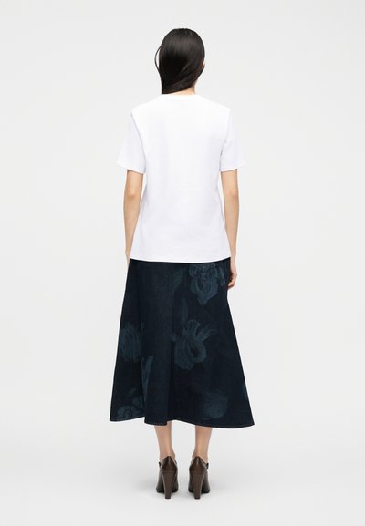 Γυναίκα με μακριά σκούρα μαλλιά που φοράει λευκό κοντομάνικο πουκάμισο, σκούρη φλοράλ μίντι φούστα και καφέ γόβες, φωτογραφημένη από πίσω.