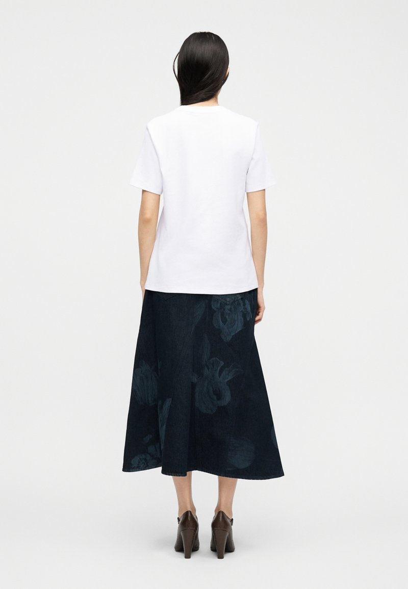 Vrouw met lang donker haar, gekleed in een witte korte mouwen overhemd, een donkere midi-rok met bloemenprint en bruine hoge hakken, gezien van achteren.