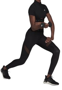 Černý sportovní outfit se skládá z přiléhavého krátkého rukávu s límečkem, legín s vysokým pasem a síťovými detaily a černých sportovních bot.