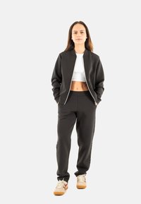 Chaqueta bomber negra con cremallera sobre una camiseta corta blanca, combinada con pantalones jogger negros. Presenta puños elásticos, un corte relajado y zapatillas deportivas.