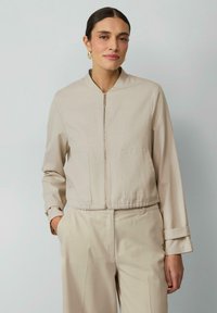 Veste beige cropped avec col montant, fermeture éclair frontale et poches plaquées. Tissu lisse avec un subtil éclat et coupe ajustée.