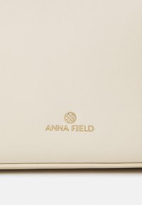 Kermanvärinen keinonahkainen laukku, jossa on kuvioitu pinta ja pohjan keskellä kohokuvioitu kultainen "ANNA FIELD" -logo.