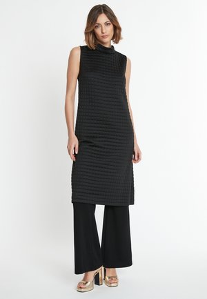 Zwart gewatteerd mouwloos jurkje met een hoge halslijn, gecombineerd met zwarte wijde broek en metallic plateau-schoenen. Gladde textuur, modern design.