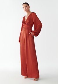 Rudos spalvos ilgos rankovės šokinėjimo kostiumas su giliu V formos kaklu ir plačiomis kelnėmis, turintis diržą ties talija ir tekstūruotą audinį.