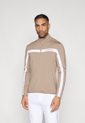 Világosbarna, cipzáras pulóver fehér csíkokkal a vállán és az ujjak mentén. Simább, rugalmas anyagból készült. Magas nyakrésszel rendelkezik.