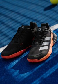 Μαύρα αθλητικά παπούτσια με υφασμάτινη άνω επιφάνεια με υφή, πορτοκαλί καουτσούκ σόλα και τρεις λευκές ρίγες στο πλάι. Τοποθετημένα σε μπλε επιφάνεια γηπέδου.