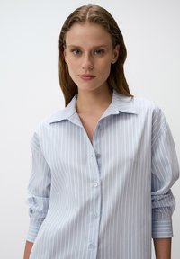 Camicia azzurra chiaro con righe bianche verticali, maniche lunghe e colletto classico. Tessuto morbido con una vestibilità rilassata.