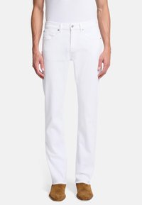 Białe dżinsy o prostym kroju nogawki, pięcio kieszeniowym stylu i z zapięciem na guzik. Model nosi beżowe zamszowe buty.