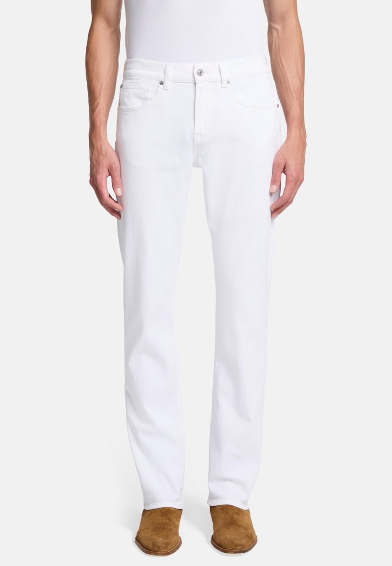 Białe dżinsy o prostym kroju nogawki, pięcio kieszeniowym stylu i z zapięciem na guzik. Model nosi beżowe zamszowe buty.