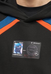 Sweat à capuche noir avec des accents bleus et orange sur les épaules, une écusson BMW M Motorsport représentant un pilote casqué, et le logo Puma Motorsport.