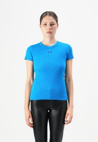 Vrouw in een aangesloten felblauw T-shirt met korte mouwen met het "HUGO" logo en zwarte leren broek, staande tegen een witte achtergrond.