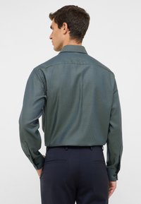 Mann trägt ein langärmeliges, gemustertes grünes Hemd, das in eine dunkelblaue Anzughose gesteckt ist. Er steht mit den Händen in den hinteren Taschen und schaut weg.