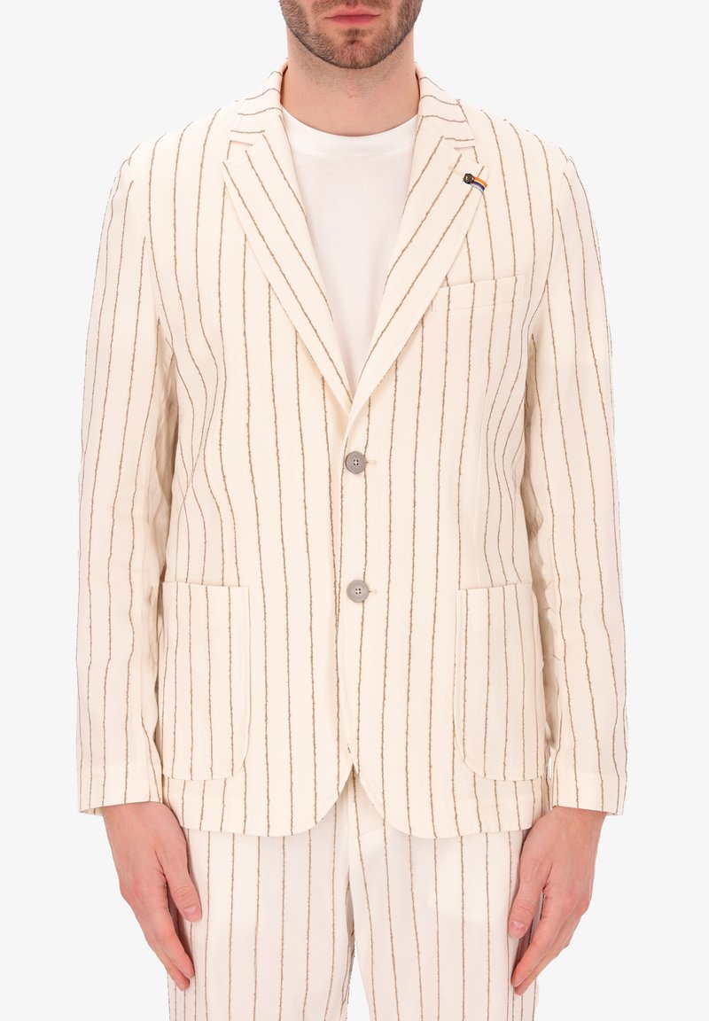 Blazer beige chiaro con righe marroni verticali, due tasche frontali, due bottoni e dettagli di cucitura discreti. Indossato sopra una t-shirt bianca.
