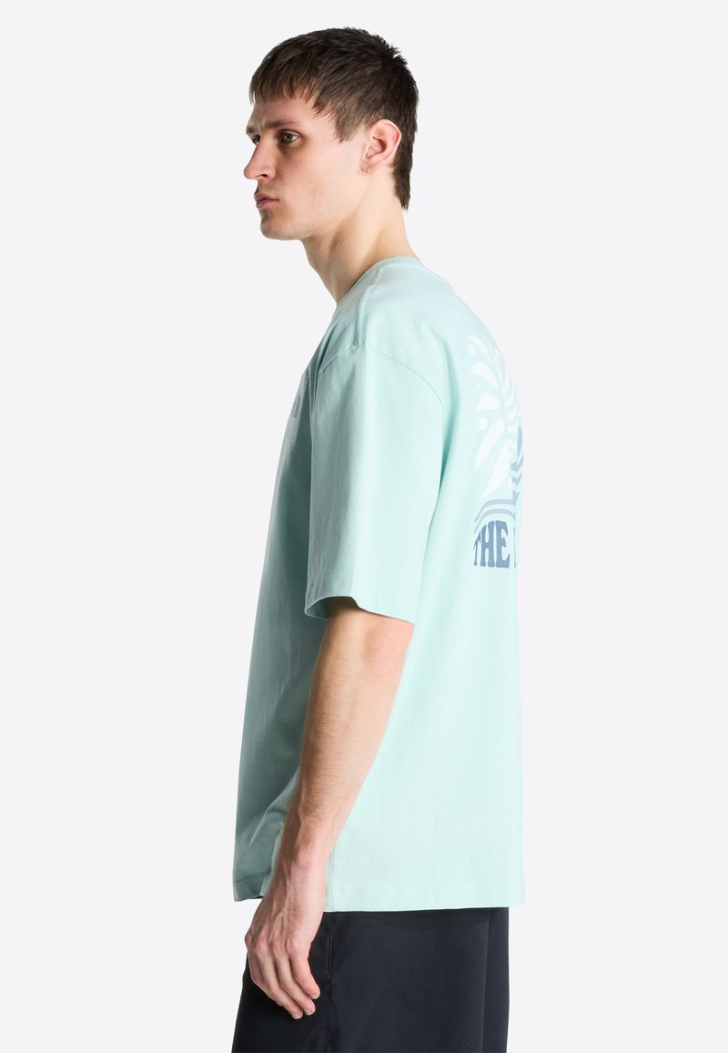 Junger Mann im mintgrünen, übergroßen T-Shirt mit Grafikdruck auf der Rückseite, stehend im Profil vor weißem Hintergrund.